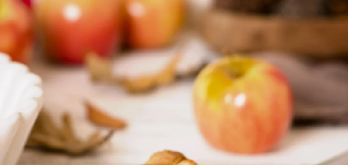 Ein Stück Apfelkuchen auf einem Kuchenheber, dekoriert mit frischen Äpfeln und Herbstblättern im Hintergrund